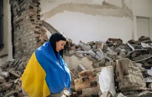 Eine in die Flagge der Ukraine gehüllte Frau steht vor den Trümmern eines zerstören Gebäudes. / Viacheslav Boiko/Shutterstock.