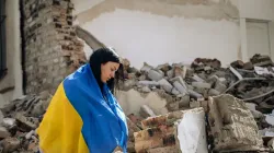 Eine in die Flagge der Ukraine gehüllte Frau steht vor den Trümmern eines zerstören Gebäudes. / Viacheslav Boiko/Shutterstock.
