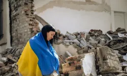 Eine in die Flagge der Ukraine gehüllte Frau steht vor den Trümmern eines zerstören Gebäudes. / Viacheslav Boiko/Shutterstock.